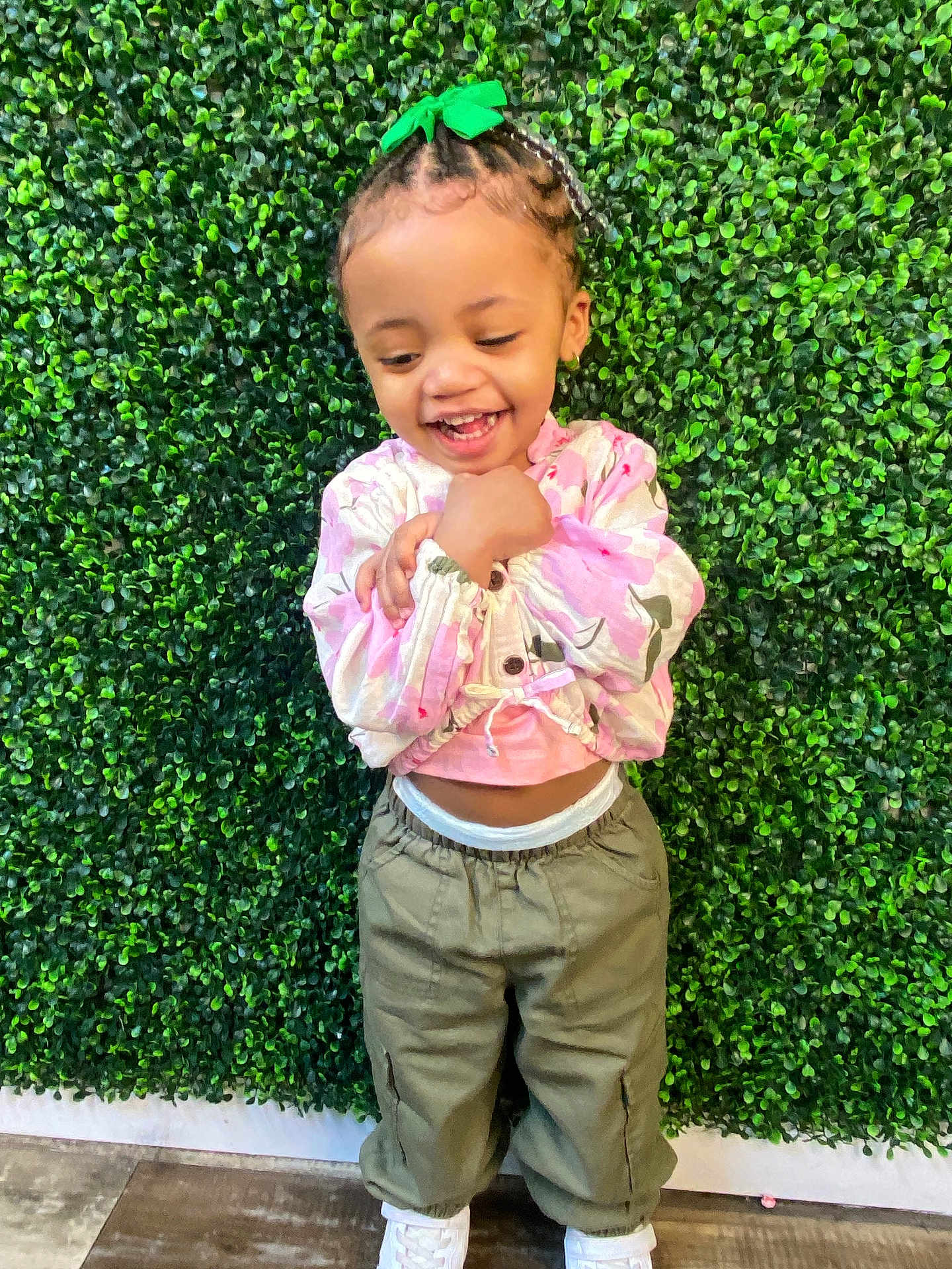 Remi is registered to the contest to win money with this photo: toddler, child, smiling, braided_hair, green_bow, pink_top, olive_pants, white_shoes, leafy_wall, green_background, standing, happy, playful, casual_clothing, young_child, outdoor_style, cute, portrait, fashion, joyful