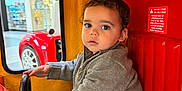 Crixus is registered to the contest to win money with this photo: child, toddler, indoor, kiddie_ride, steering_wheel, curly_hair, gray_sweater, camouflage_pants, red_seat, yellow_wall, toy_car, window, bracelet, necklace, face, eyes, hand, seat, play, vehicle