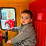 child, toddler, indoor, kiddie_ride, steering_wheel, curly_hair, gray_sweater, camouflage_pants, red_seat, yellow_wall, toy_car, window, bracelet, necklace, face, eyes, hand, seat, play, vehicle