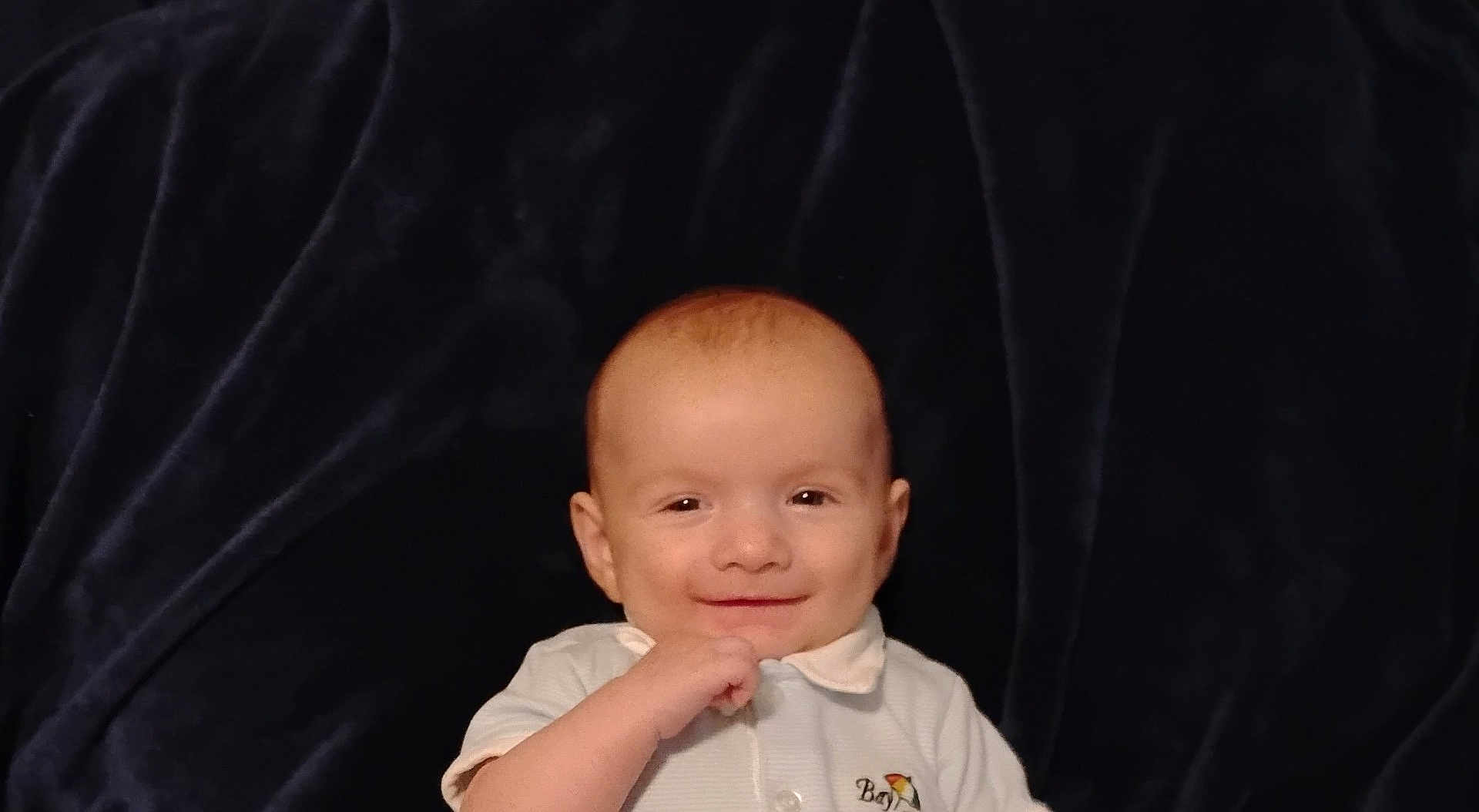 Jaxon joined the competition — help win amazing prizes! baby, smiling, infant, child, white_shirt, collar, hand, face, head, skin, lying_down, blanket, dark_background, portrait, cute, happy, young, person, indoors, closeup