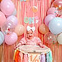 Emma is registered to the contest to win money with this photo: baby, high_chair, balloons, party, celebration, pink, pastel_colors, tassels, decorations, smiling, child, headband, white_bow, festive, milestone, indoor, happy, cute, infant, birthday