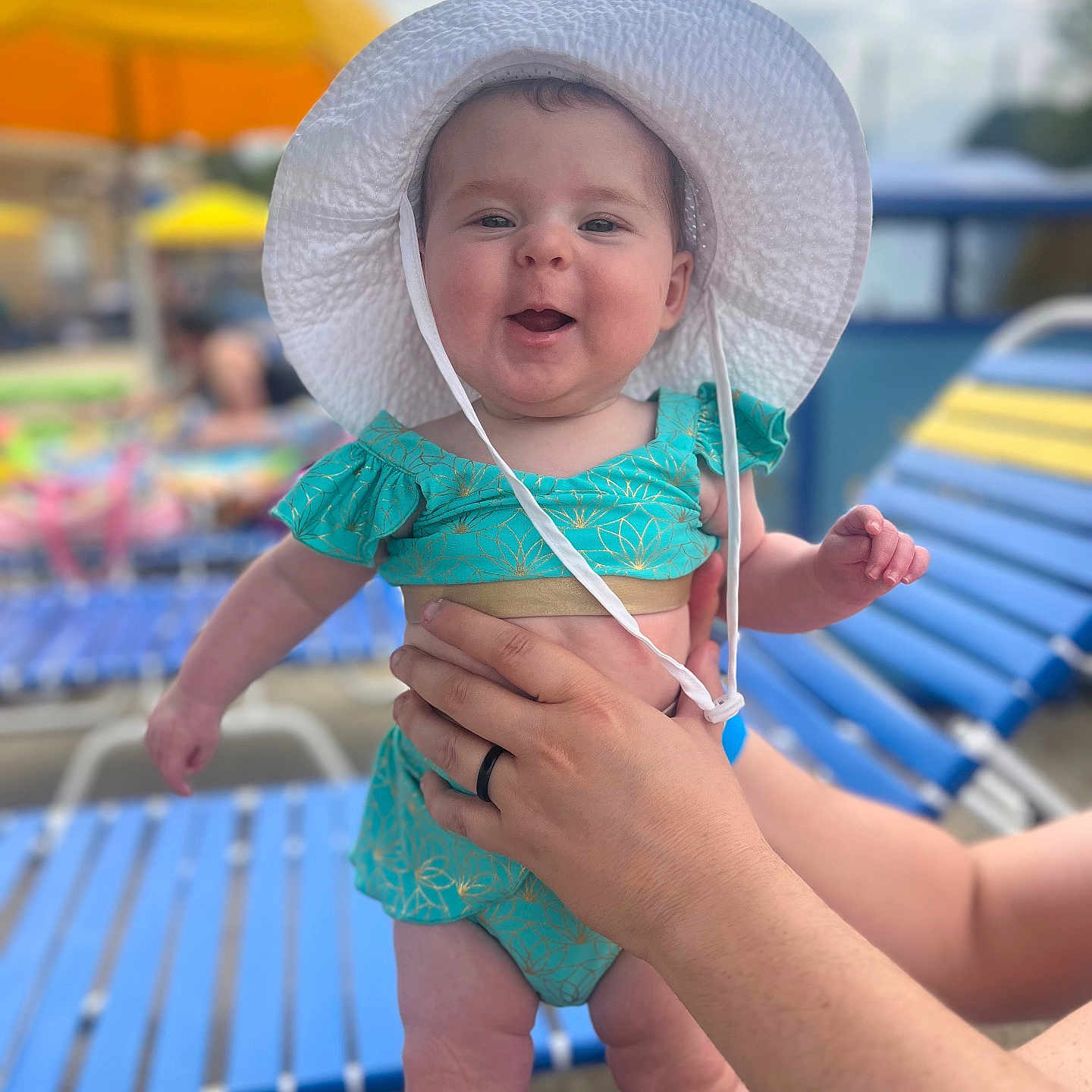 Sutton is registered to the contest to win money with this photo: baby, child, smiling, white_hat, turquoise_outfit, hands, adult, poolside, lounge_chair, yellow_umbrella, summer, outdoor, sunny, happy, person, infant, cute, vacation, bright, daytime