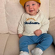 Hudson joined the competition — help win amazing prizes! baby, child, cap, yellow_hat, white_sweater, embroidered_sweater, blue_pants, white_sneakers, couch, smile, happy, indoor, portrait, sitting, cute, fashion, footwear, headwear, young_child, cozy