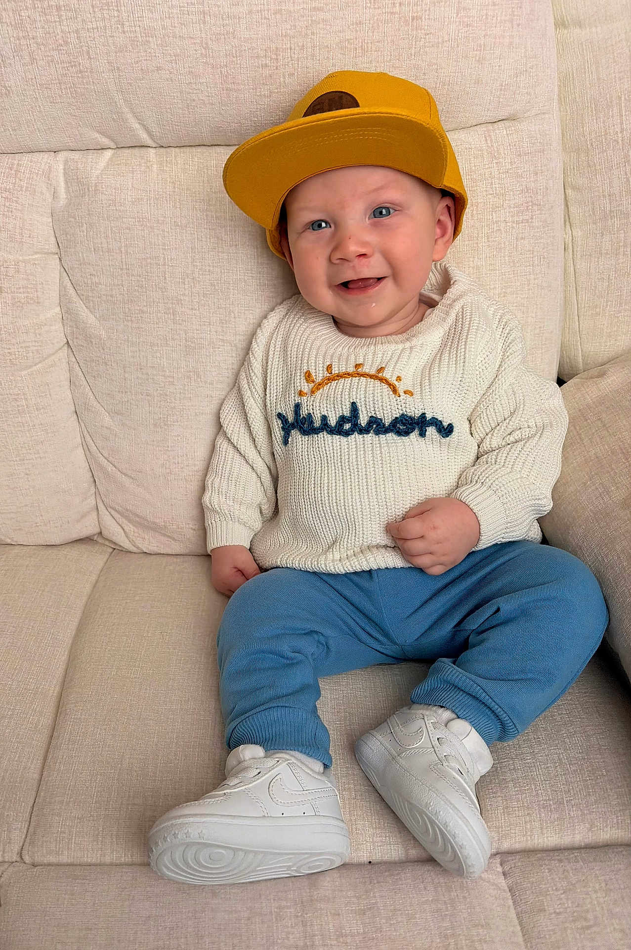 Hudson joined the competition — help win amazing prizes! baby, child, cap, yellow_hat, white_sweater, embroidered_sweater, blue_pants, white_sneakers, couch, smile, happy, indoor, portrait, sitting, cute, fashion, footwear, headwear, young_child, cozy
