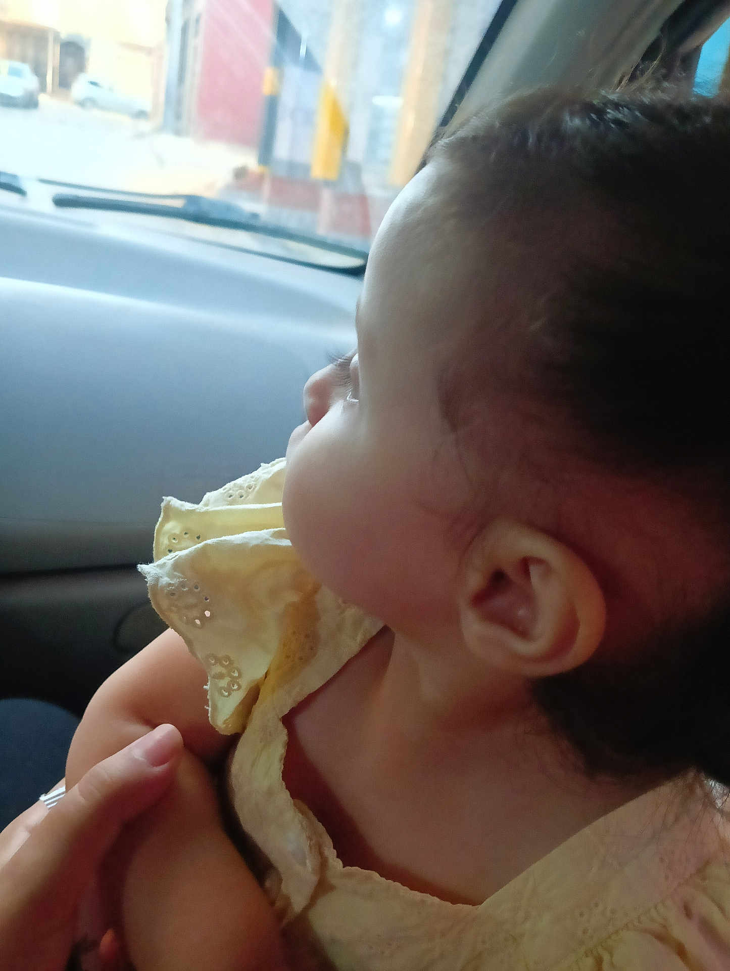 Farah is registered to the contest to win money with this photo: baby, child, side_profile, yellow_dress, car_interior, hand, person, curious, infant, skin, ear, face, window, seat, vehicle, portrait, young_child, holding, clothing, close_up