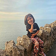 Hadia is registered to the contest to win money with this photo: black_tshirt, camouflage_pants, casual_clothing, child, cloudy_sky, coast, daylight, happy, nature, ocean, outdoor, portrait, posing, rocks, sandals, scenery, sea, smiling, sunset, young