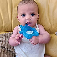 Andréa participe au concours pour gagner de l'argent avec cette photo : baby, blue_eyes, teether, white_outfit, yellow_couch, blanket, infant, cute, child, sitting, indoors, portrait, skin, hand, face, mouth, toy, comfort, young, expression