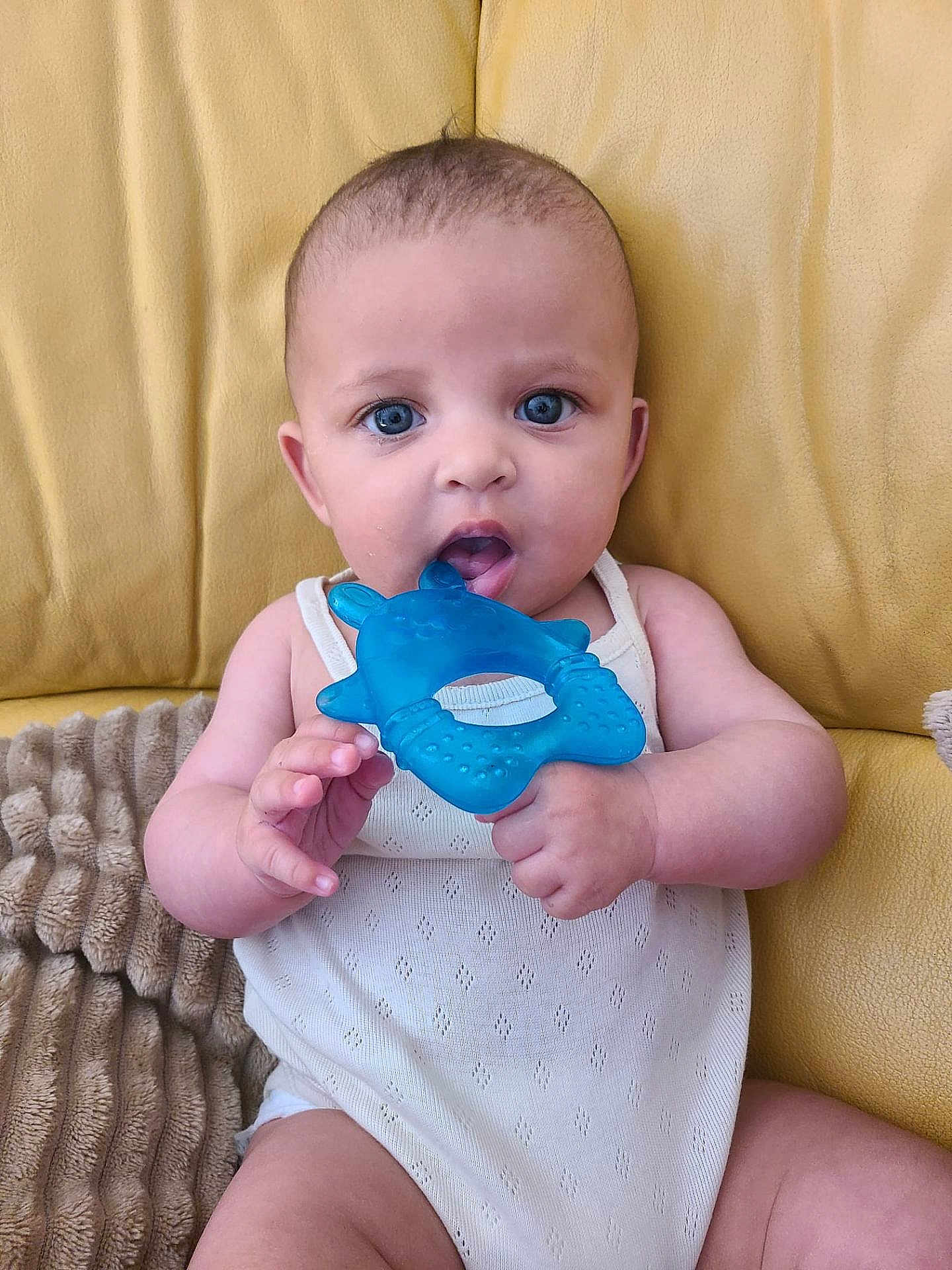 Andréa participe au concours pour gagner de l'argent avec cette photo : baby, blue_eyes, teether, white_outfit, yellow_couch, blanket, infant, cute, child, sitting, indoors, portrait, skin, hand, face, mouth, toy, comfort, young, expression
