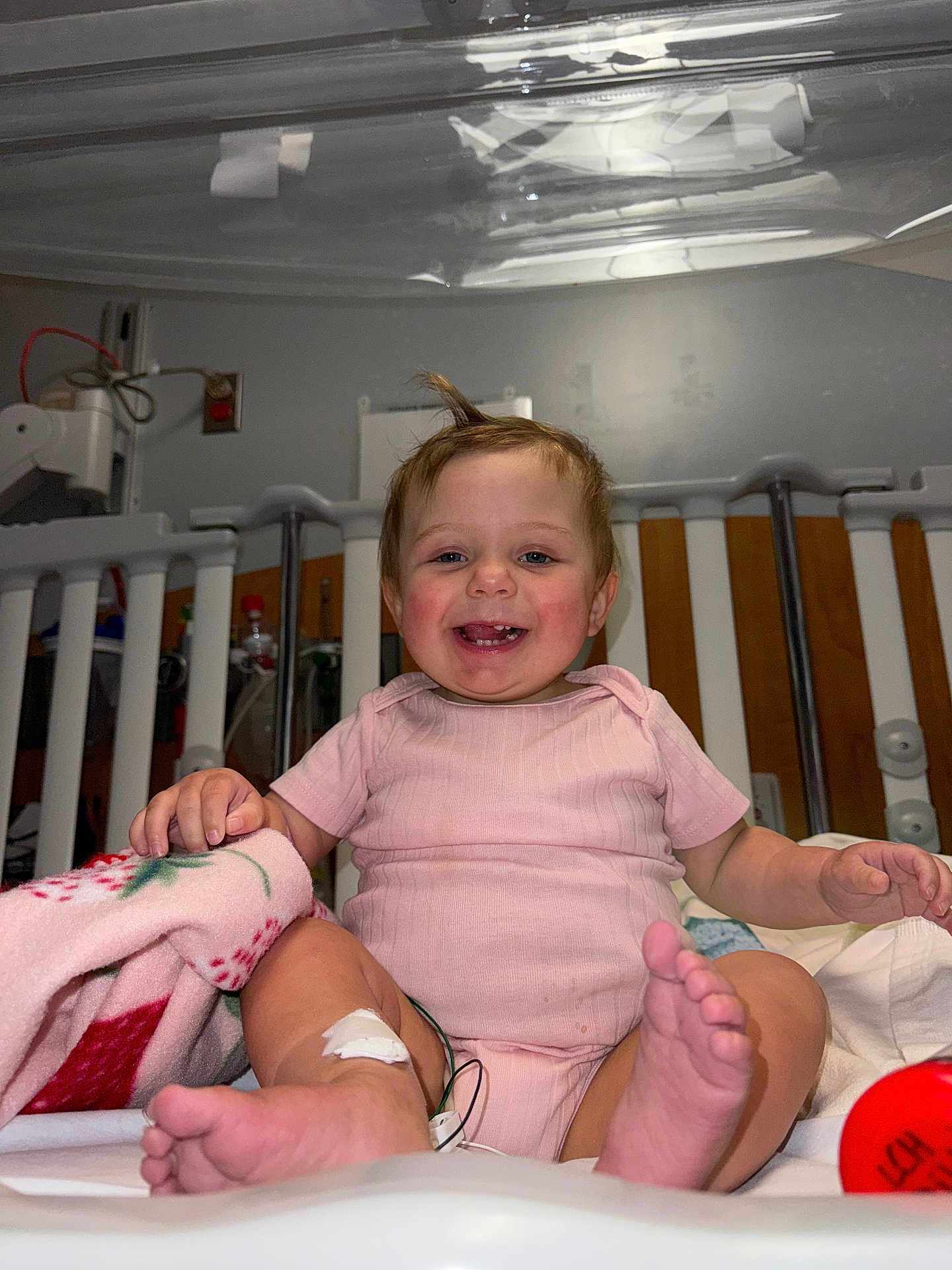 Paisley is registered to the contest to win money with this photo: baby, child, smiling, hospital_crib, pink_onesie, medical_tape, feet, hand, blanket, hair, toddler, indoor, happy, face, person, crib_bars, toy, red_object, skin, cute