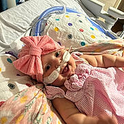 Paisley joined the competition — help win amazing prizes! baby, child, pink_bow, nasal_cannula, hospital_bed, pillow, blanket, smiling, infant, cute, medical_equipment, person, indoors, clothing, headwear, toddler, happy, lying_down, healthcare, soft_lighting