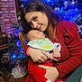 woman, baby, christmas_tree, holiday, festive_clothing, red_sweater, sleeping_baby, indoor, decorations, lights, ornaments, love, family, holding, smile, chair, wood_floor, bracelet, bib, peaceful