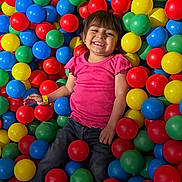 Blanca is registered to the contest to win money with this photo: child, girl, smiling, happy, ball_pit, colorful, plastic_balls, playground, indoor, portrait, sitting, pink_shirt, jeans, brown_hair, bangs, face, teeth, wristband, playful, recreation