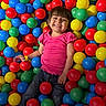 child, girl, smiling, happy, ball_pit, colorful, plastic_balls, playground, indoor, portrait, sitting, pink_shirt, jeans, brown_hair, bangs, face, teeth, wristband, playful, recreation