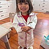 child, toddler, girl, smiling, portrait, standing, indoors, kitchen, white_coat, costume, pretend_play, toy, purple_object, slippers, wooden_floor, cabinets, high_chair, home, happy, face