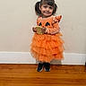 child, girl, toddler, portrait, smiling, halloween, costume, pumpkin, orange_dress, ruffles, waffle, food, pigtails, indoors, standing, wooden_floor, wall, sneakers, happy, face