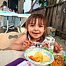 child, toddler, girl, smiling, happy, portrait, outdoor, backyard, table, food, snacks, chips, juice_box, plate, high_chair, pool, fence, tree, family, daylight