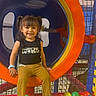 child, girl, toddler, smiling, portrait, indoor, playground, play_tube, play_structure, climbing_net, ball_pit, colorful, orange, yellow, blue, seated, play_area, casual_clothing, pigtails, happy