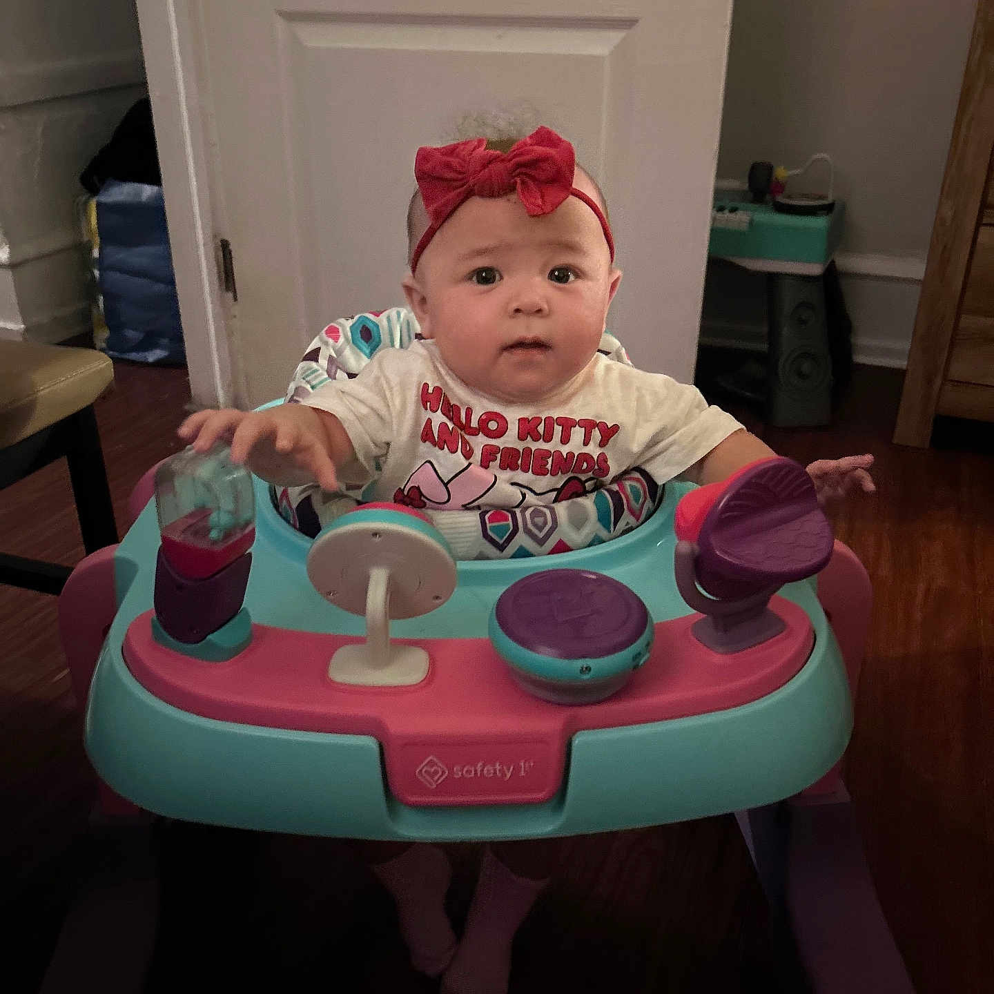 Armani is registered to the contest to win money with this photo: baby, child, infant, headband, red_bow, hello_kitty, shirt, baby_walker, toy, indoor, door, floor, wood_floor, furniture, chair, curious, person, caucasian, expression, sitting