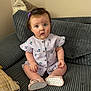 baby, child, infant, sitting, purple_bow, dress, cherry_pattern, couch, gray_couch, pillow, footwear, shoes, heart_pattern, indoors, curious_expression, plush, soft_lighting, cute, person, young_child