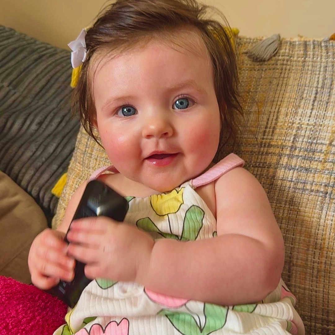 Aoibhí joined the competition — help win amazing prizes! baby, blue_eyes, bow, child, chubby_cheeks, couch, cozy, cute, flower_dress, green, hair_accessory, indoor, person, pink, portrait, remote_control, sitting, smiling, soft_lighting, yellow