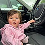 baby, child, car_interior, bmw, steering_wheel, pink_hoodie, socks, seat, window, side_mirror, curious_expression, blue_eyes, short_hair, car_seat, dashboard, overhead_light, gray_seat, hand_on_steering_wheel, vehicle, daytime