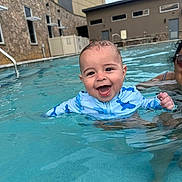 Jettson is registered to the contest to win money with this photo: baby, child, pool, water, swimming, smiling, happy, person, adult, building, stone_wall, swim_shirt, blue, outdoor, summer, splashing, face, fun, leisure, sunlight