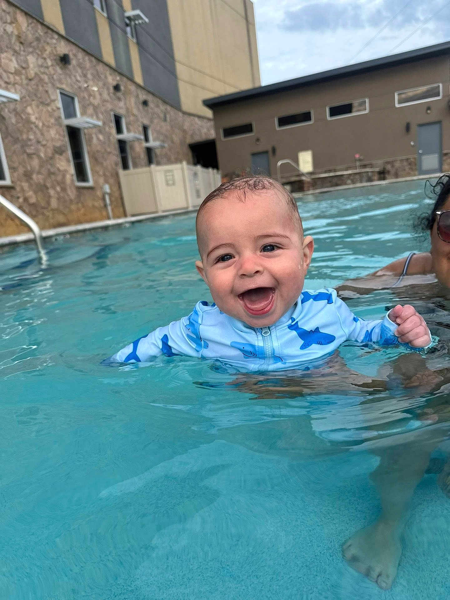 Jettson is registered to the contest to win money with this photo: baby, child, pool, water, swimming, smiling, happy, person, adult, building, stone_wall, swim_shirt, blue, outdoor, summer, splashing, face, fun, leisure, sunlight