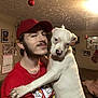 man, dog, white_dog, red_cap, red_shirt, indoor, living_room, holiday_decorations, christmas_ornaments, stocking, beard, chain_necklace, calendar, couch, blanket, picture_frames, pet, holding, smile, casual_clothing
