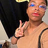 person, glasses, peace_sign, cat, indoor, floor, curtain, tshirt, lipstick, hand, brown_skin, short_hair, watch, ring, selfie, casual, background, text_overlay, blue_reflection, brown_cat