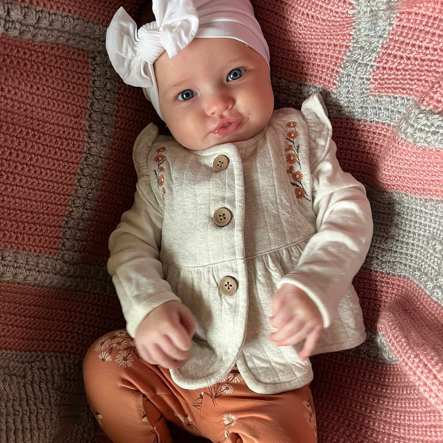 Kymber is registered to the contest to win money with this photo: baby, blanket, blue_eyes, bow, child, cozy, cute, floral_embroidery, gray_blanket, headband, indoors, infant, knitted_blanket, leggings, orange_leggings, person, pink_blanket, portrait, sitting, vest