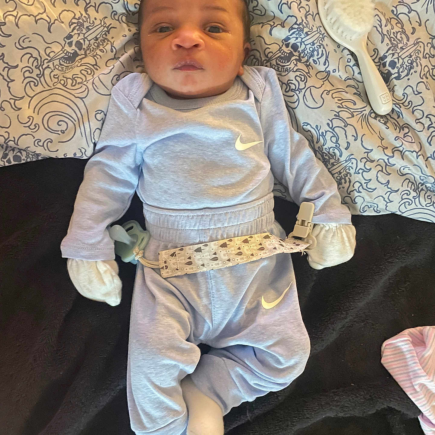 Tristan is registered to the contest to win money with this photo: baby, infant, child, portrait, lying_down, indoors, blanket, patterned_blanket, gray_outfit, sportswear, swoosh_logo, mittens, pacifier, pacifier_clip, hairbrush, bed, textile, sleepy, looking_at_camera, cozy