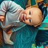 baby, infant, child, portrait, smiling, lying_down, blanket, shaggy_blanket, white_clothing, blue_eyes, bald, toy, colorful_toy, nursery, indoor, soft_texture, cozy, close_up, happy, photography