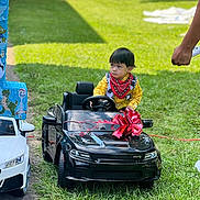 Aiden is registered to the contest to win money with this photo: child, toy_car, red_bow, yellow_shirt, bandana, outdoor, grass, sidewalk, hand, remote_control, trees, daylight, costume, vehicle, playing, portrait, person, black_car, white_car, decoration