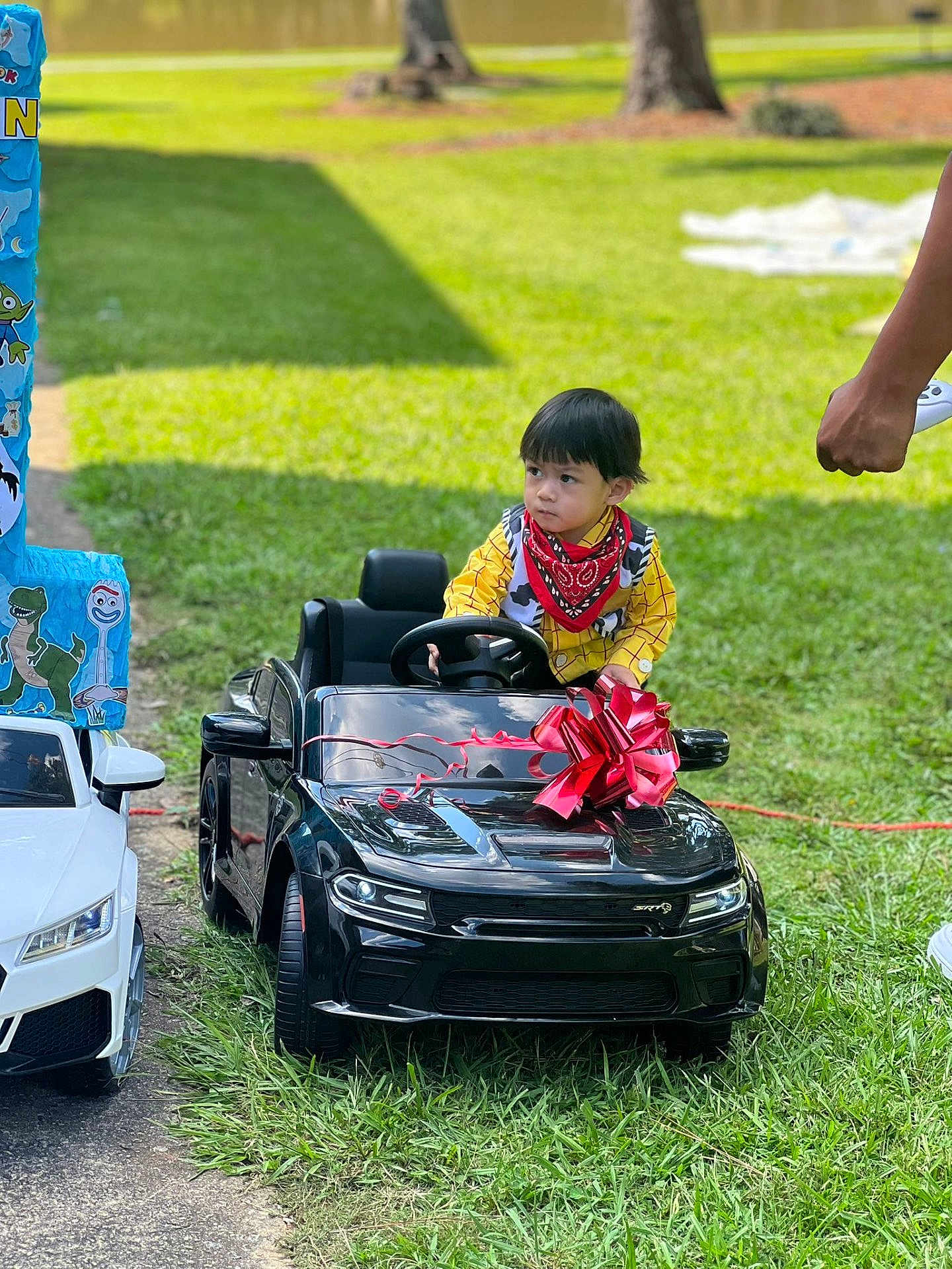 Aiden is registered to the contest to win money with this photo: child, toy_car, red_bow, yellow_shirt, bandana, outdoor, grass, sidewalk, hand, remote_control, trees, daylight, costume, vehicle, playing, portrait, person, black_car, white_car, decoration