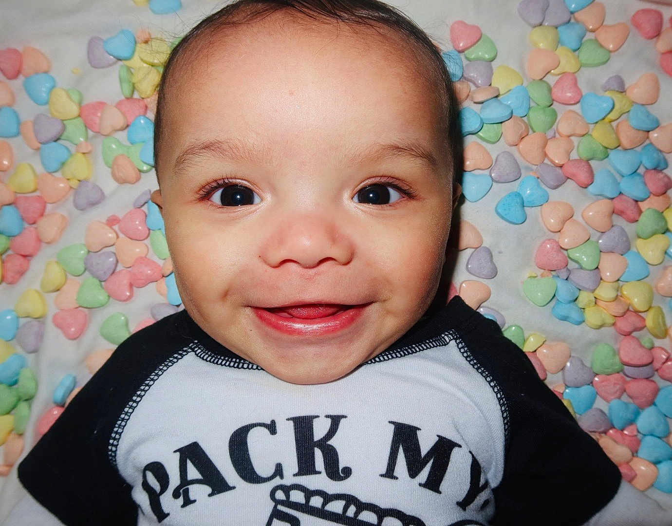 Abdul-Malik is registered to the contest to win money with this photo: baby, infant, smiling, closeup, face, big_eyes, dimples, cheeks, onesie, clothing, pastel, candy_hearts, heart_shapes, background, portrait, happy, lying_down, skin, cute, playful