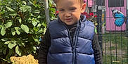 Liam a rejoint le concours — aidez-le/la à gagner de superbes lots ! toddler, child, blue_vest, jeans, sneakers, gravel, garden, flowers, plants, butterfly, pink_door, outdoor, daylight, fence, greenery, casual_clothing, sunlight, portrait, cute, young_child