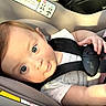 baby, infant, child, car_seat, car, safety_harness, seatbelt, portrait, face, eyes, looking_at_camera, wide_eyes, brown_hair, smiling, pink_clothing, indoor, vehicle, travel, sunlight, close_up