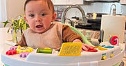 Hiraya Wynne joined the competition — help win amazing prizes! baby, child, infant, kitchen, activity_walker, toy, sitting, face, portrait, wide_eyes, open_mouth, brown_hair, bib, clothing, bare_feet, furniture, cabinets, sink, microwave, houseplant