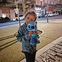 child, plush_toy, denim_jacket, sidewalk, night, street, cars, urban, building, tree, toy, cute, outdoor, person, holding, blue, casual_clothing, happy, playful, blurry