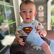 Walker is registered to the contest to win money with this photo: baby, child, superman, overalls, saddle, hand, portrait, blue_eyes, outdoor, daylight, curious, person, cute, short_hair, decorative, clothing, holding, seated, expression, window