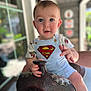 baby, child, superman, overalls, saddle, hand, portrait, blue_eyes, outdoor, daylight, curious, person, cute, short_hair, decorative, clothing, holding, seated, expression, window