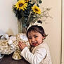 toddler, child, toy, plush_giraffe, table, sunflowers, flowers, hair_bows, smile, indoor, cute, happy, knit_vest, white_shirt, person, face, earrings, wood, decor, nature