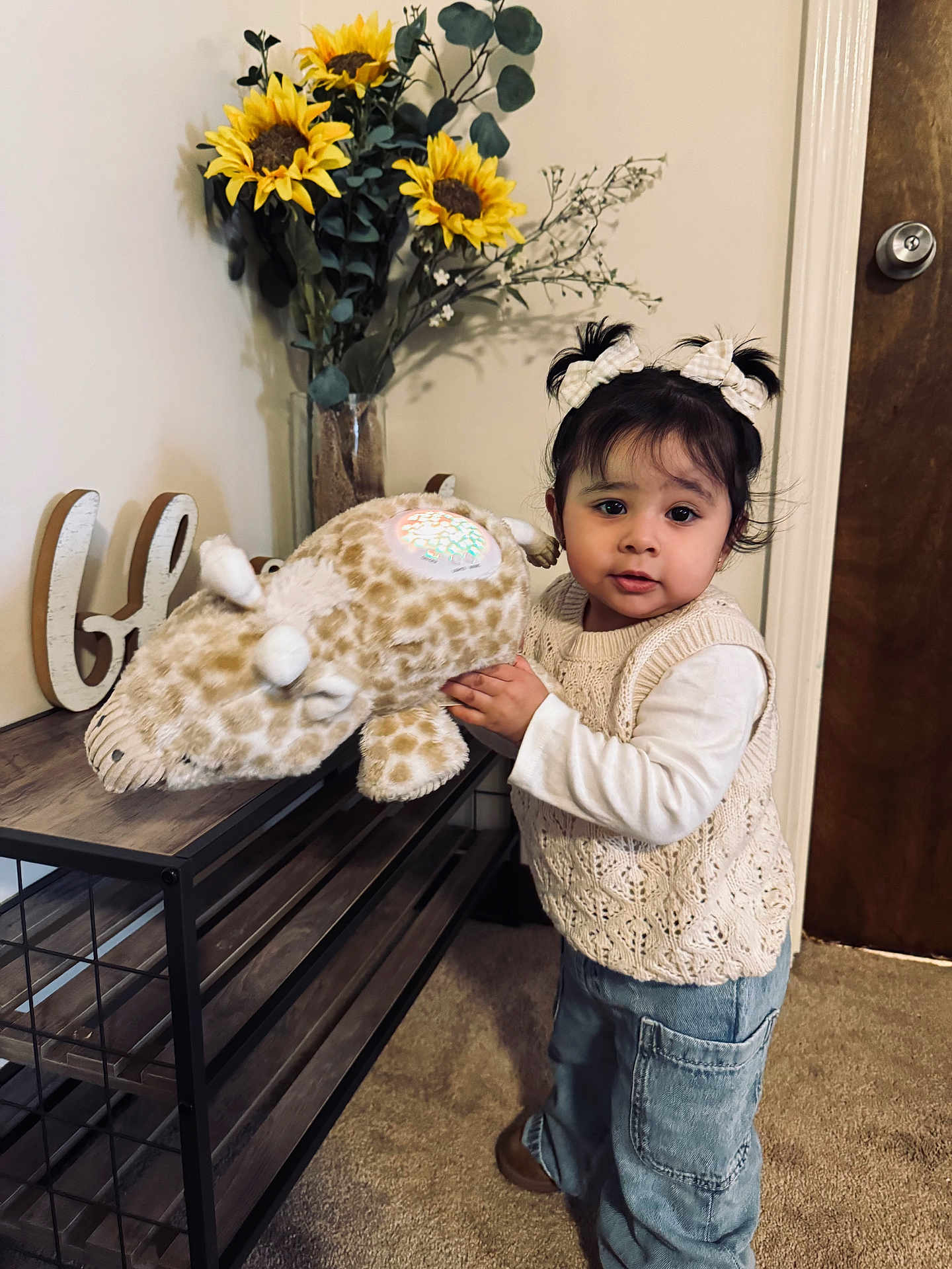 Luna joined the competition — help win amazing prizes! toddler, child, indoor, plush_toy, giraffe, flower_vase, sunflower, carpet, clothing, pigtails, hair_bows, table, door, cute, person, young_child, knit_sweater, jeans, toy, home_decor