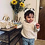 toddler, child, girl, smile, indoor, clothing, jeans, sweater, hair_bows, toy, plush_giraffe, furniture, flowers, sunflowers, vase, carpet, door, wooden_door, shelf, happy