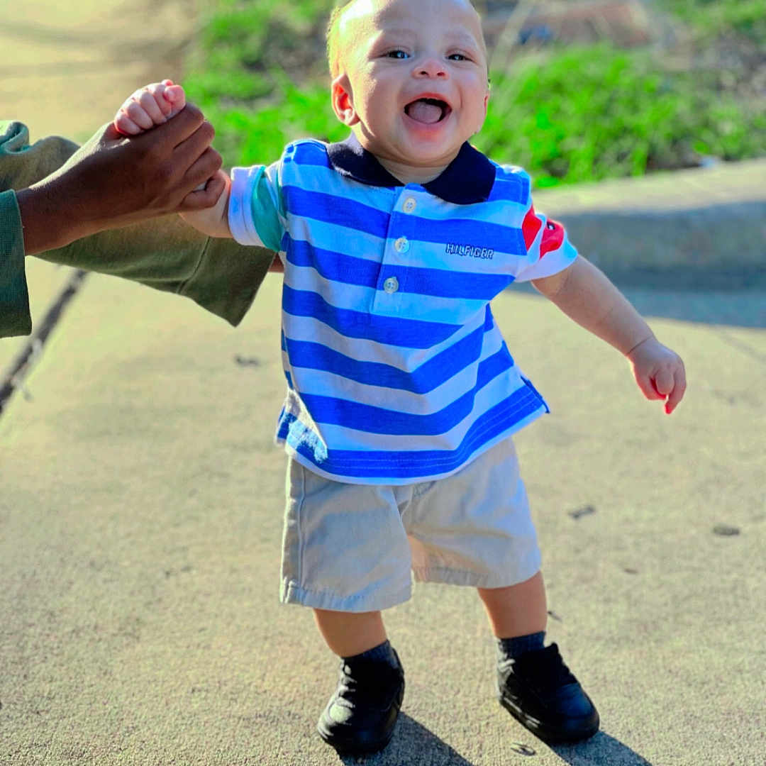 Deontae is registered to the contest to win money with this photo: blue_shirt, casual_clothing, child, daylight, grass, happy, holding_hand, nature, outdoor, person, shadow, shoes, shorts, sidewalk, smiling, striped_shirt, sunlight, support, toddler, walking