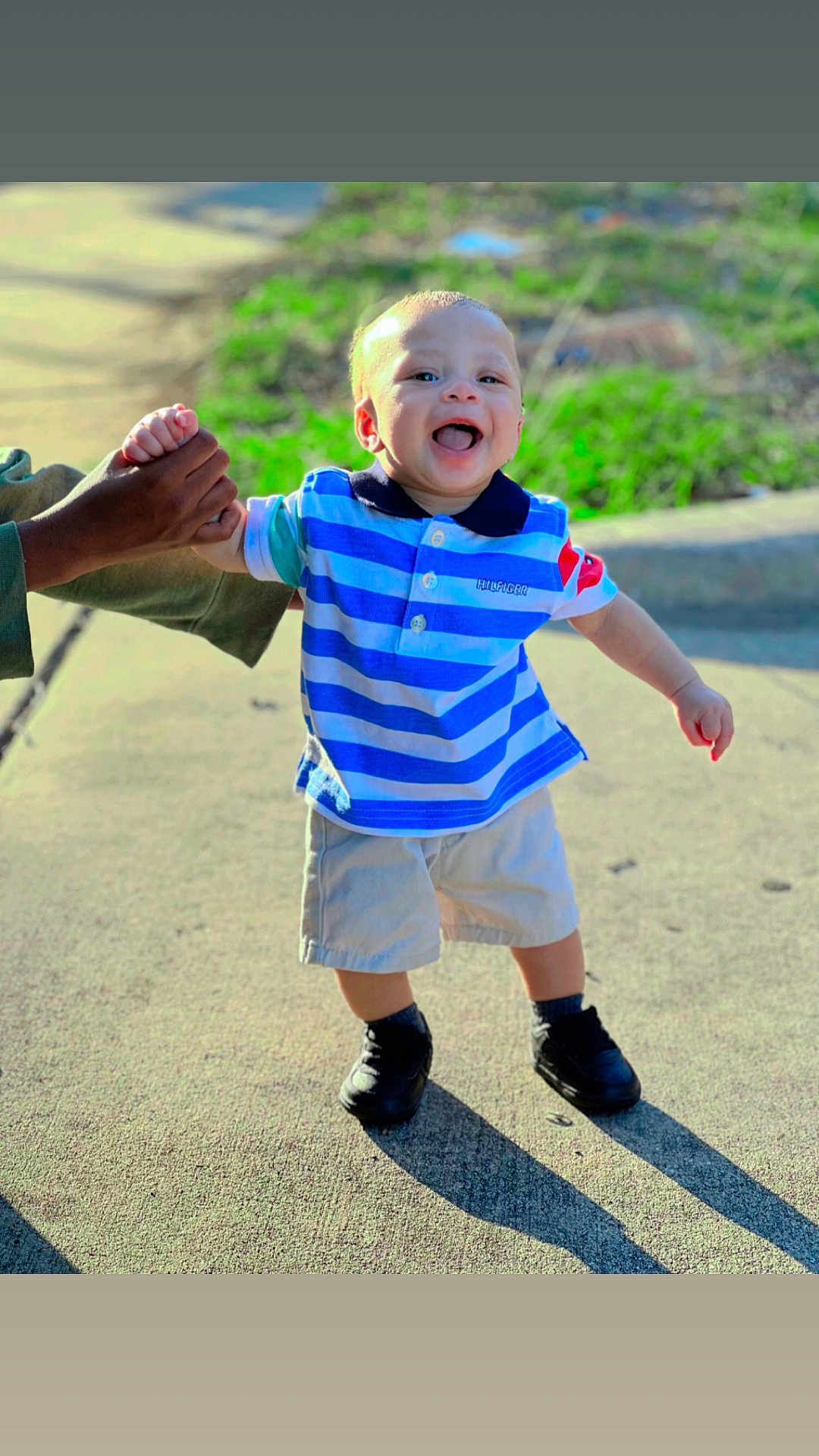 Deontae is registered to the contest to win money with this photo: child, toddler, smiling, walking, outdoor, sidewalk, holding_hand, sunlight, shadow, blue_shirt, striped_shirt, shorts, shoes, happy, person, daylight, nature, grass, casual_clothing, support