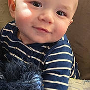 Logan joined the competition — help win amazing prizes! baby, child, striped_clothing, plush_toy, teddy_bear, couch, blanket, dinosaur_pattern, portrait, smiling, indoor, cozy, cute, young_child, face, head, soft_toy, seated, striped_shirt, happy