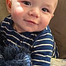 baby, child, striped_clothing, plush_toy, teddy_bear, couch, blanket, dinosaur_pattern, portrait, smiling, indoor, cozy, cute, young_child, face, head, soft_toy, seated, striped_shirt, happy