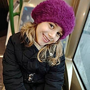 Gayané a rejoint le concours — aidez-le/la à gagner de superbes lots ! black_coat, blonde_hair, casual, child, closeup, cold_weather, face, girl, happy, hat, indoor, natural_light, person, portrait, purple_hat, sitting, smiling, warm_clothing, window, winter_coat