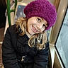 black_coat, blonde_hair, casual, child, closeup, cold_weather, face, girl, happy, hat, indoor, natural_light, person, portrait, purple_hat, sitting, smiling, warm_clothing, window, winter_coat